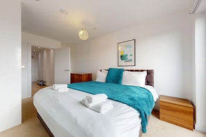 A modern bedroom with a large bed, a wooden nightstand, a wall-mounted artwork, and a door leading to another room. - 1BR Flat w/Balcony Near Tube and London Stadium (London)