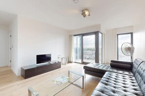 A modern living room with a flat-screen TV, a glass coffee table, a leather sofa, and a large sliding glass door leading to a balcony. - 1BR Flat w/Balcony Near Tube and London Stadium (London)
