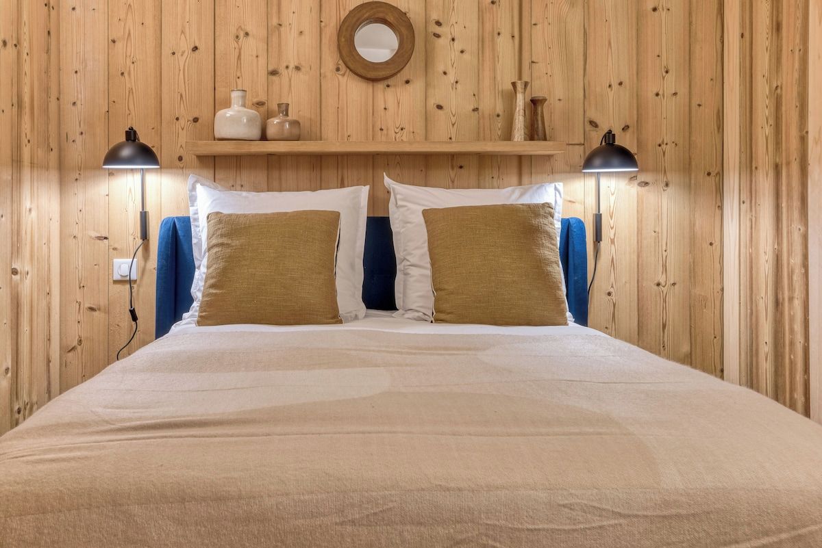 Un lit avec une tête de lit en bois, deux lampes de chevet et une étagère ornée d’objets décoratifs.