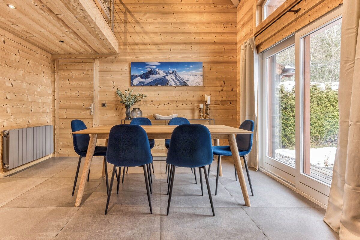 Un interior de madera con una mesa de comedor y sillas, un cuadro de una montaña y una ventana con vistas a árboles.