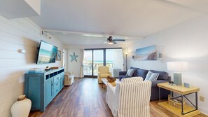 Ein geräumiges Wohnzimmer mit Fernseher, Esstisch und einer Couch. Ein Balkon bietet Blick auf den Strand.