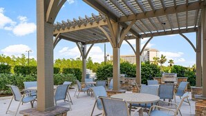 An outdoor patio area with tables and chairs, a pergola structure, and a stone fireplace. - Luau 2 6622/6624LO, New to Rentals, Pool Views, (Miramar Beach)