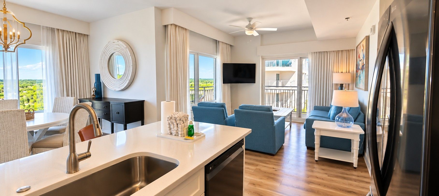 A modern hotel room with a kitchen area, a seating area with blue sofas, a balcony with a view, and a flat-screen TV mounted on the wall.