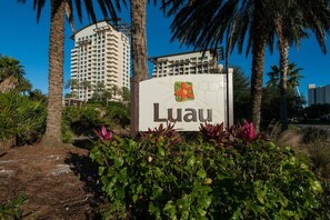 A sign with the name 'Luau' and a logo, surrounded by greenery and palm trees. - New to Rental Program | Luau Lagoon Hideaway | Two Queen Beds (Miramar Beach)