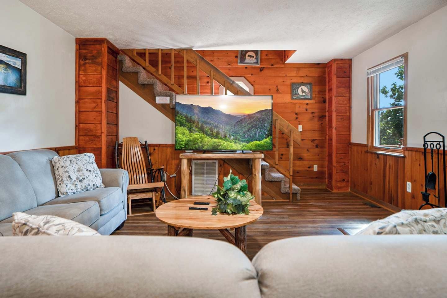 A cozy living room with a wooden staircase, a flat-screen TV on a wooden stand, a grey sofa, and a round coffee table with a plant.