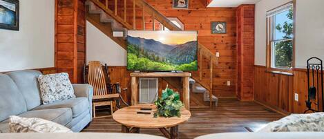 A cozy living room with a wooden staircase, a flat-screen TV on a wooden stand, a grey sofa, and a round coffee table with a plant.