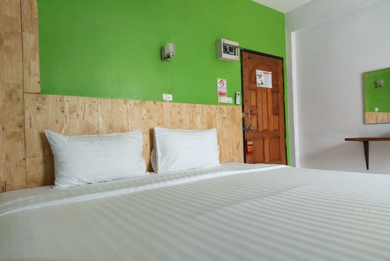A hotel room with a wooden headboard, a bed with white bedding, a green wall, a wooden door, and a small table with a mirror.