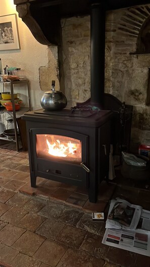 A wood-burning stove with a visible fire, a kettle on top, and a brick floor. - Cottage 'La Maison Des  Beaux Jours' with Private Terrace, Private Garden and Wi-Fi (Montaron)