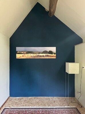 A room with a dark blue wall, a framed picture of a field with animals, a white lamp, and a patterned rug. - Cottage 'La Maison Des  Beaux Jours' with Private Terrace, Private Garden and Wi-Fi (Montaron)