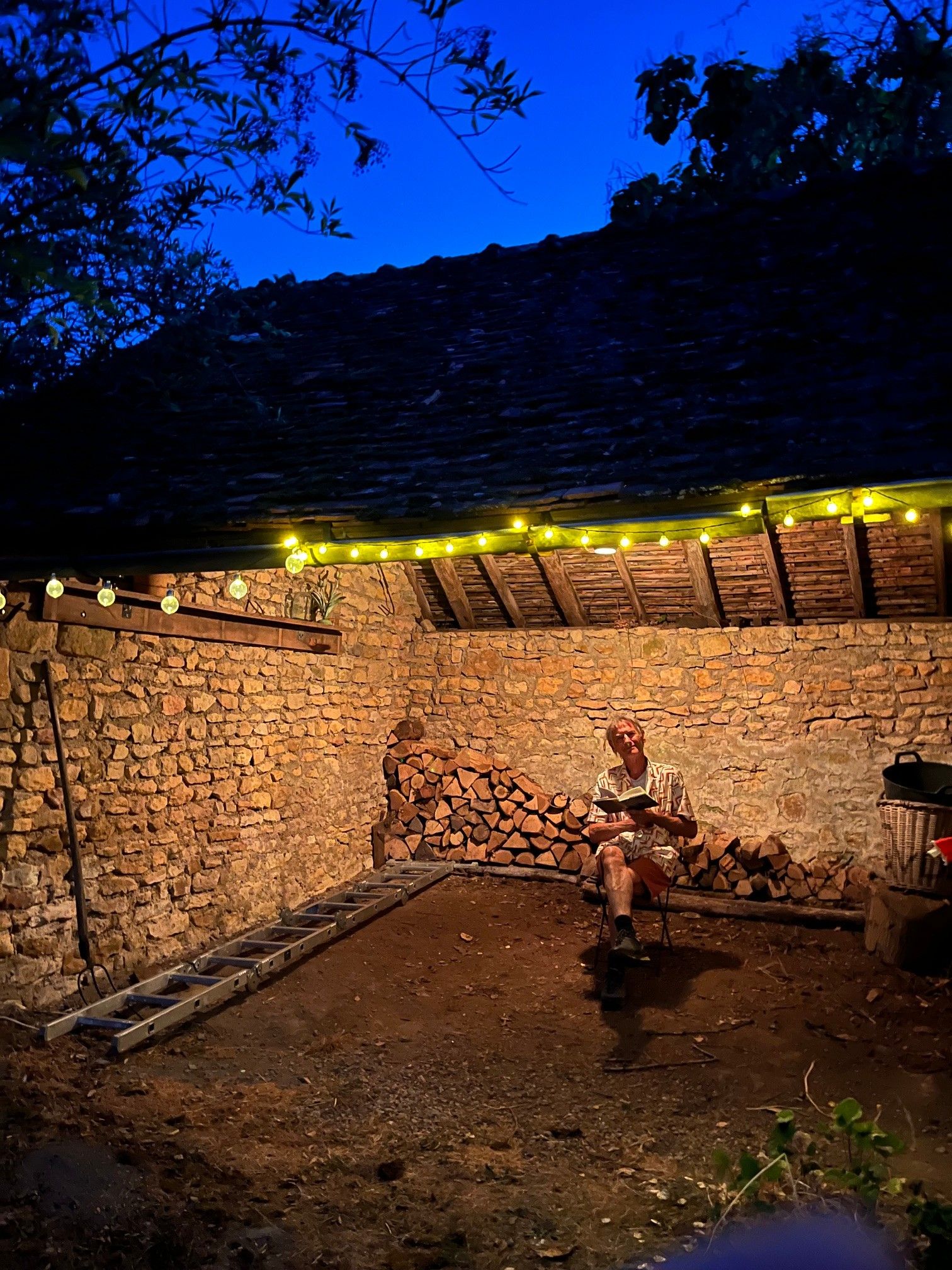 Une personne est assise sur une chaise dans un espace extérieur dont les murs sont en pierre, avec un plafond en bois et des guirlandes lumineuses.