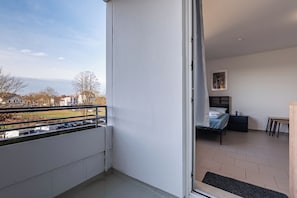 A balcony with a view of a parking area and buildings, a bed with a headboard, a small table, and a chair.