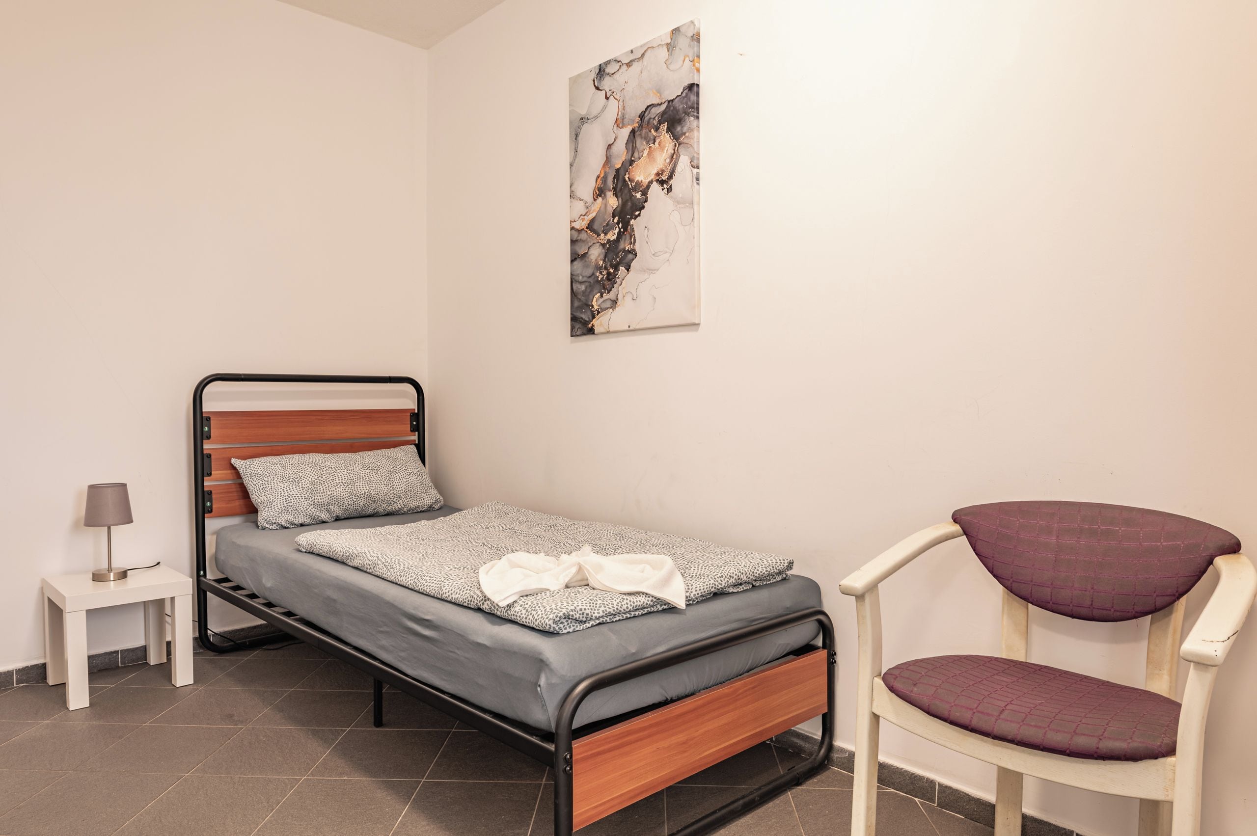 Un lit simple avec une tête de lit en bois, une petite table de chevet avec une lampe, une chaise avec un siège rembourré et un tableau abstrait accroché au mur.