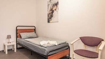 A single bed with a wooden headboard, a small bedside table with a lamp, a chair with a cushioned seat, and a wall-mounted abstract painting.