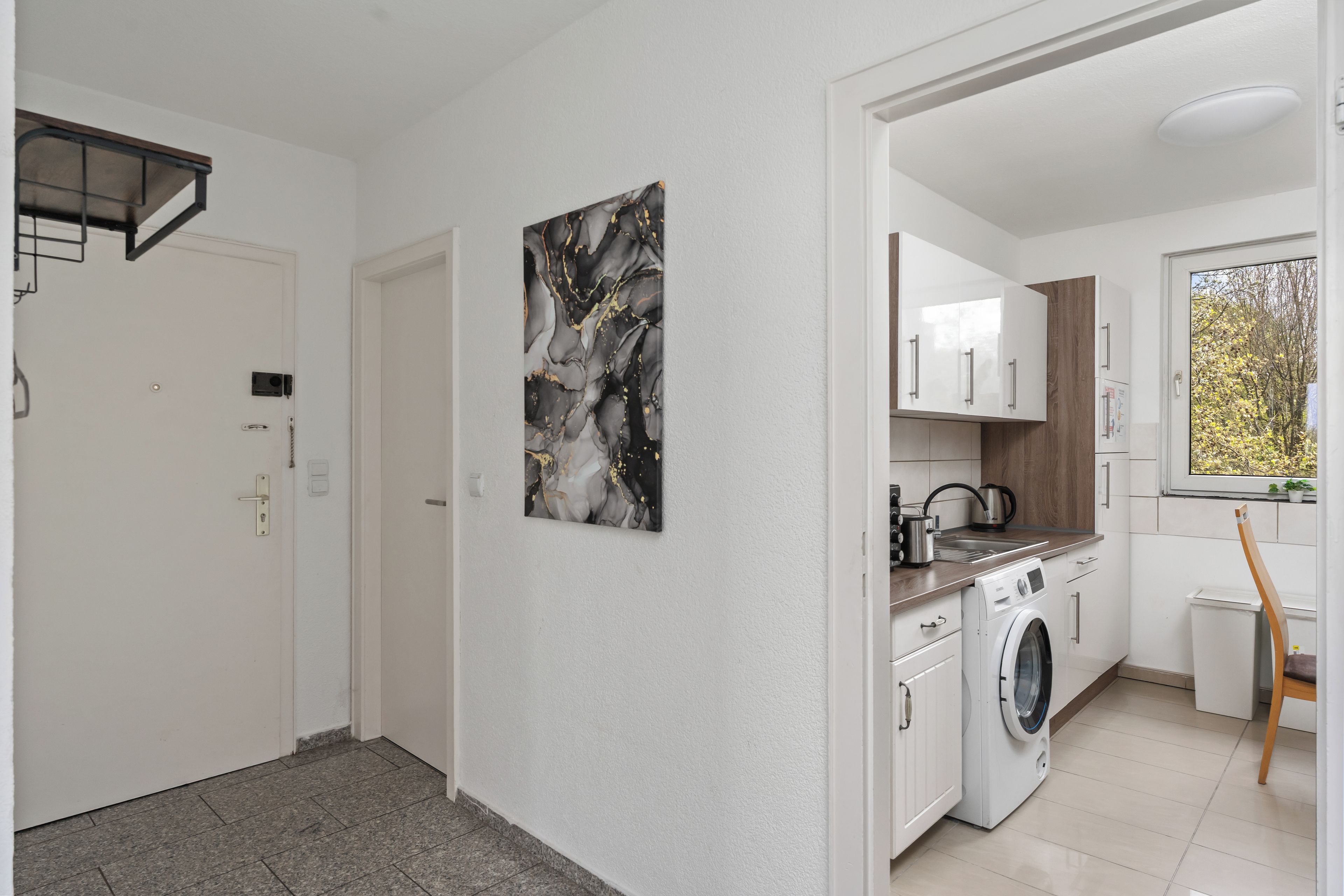 A modern kitchen with a built-in washing machine, a window with a view of trees, and a marble artwork on the wall.