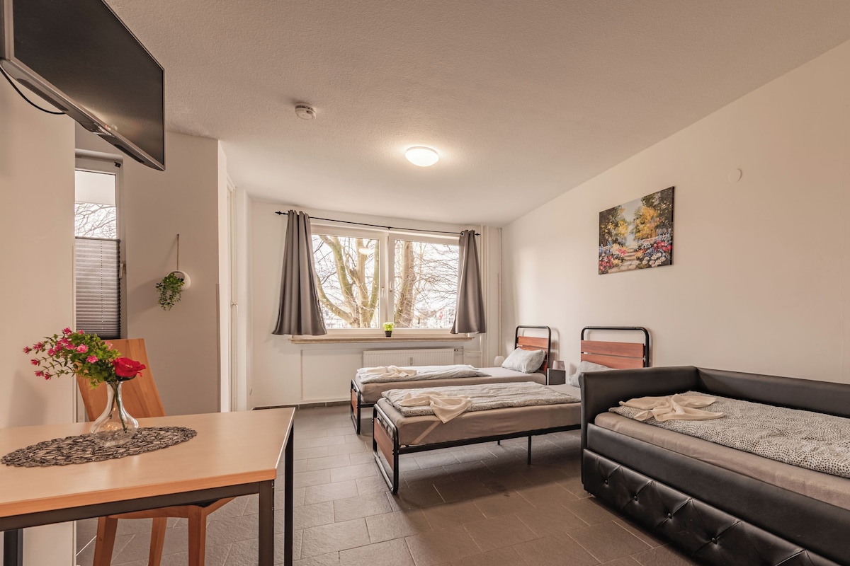Habitació compacta amb dos llits individuals, un sofà, una taula amb un ram de flors i una finestra amb cortines.