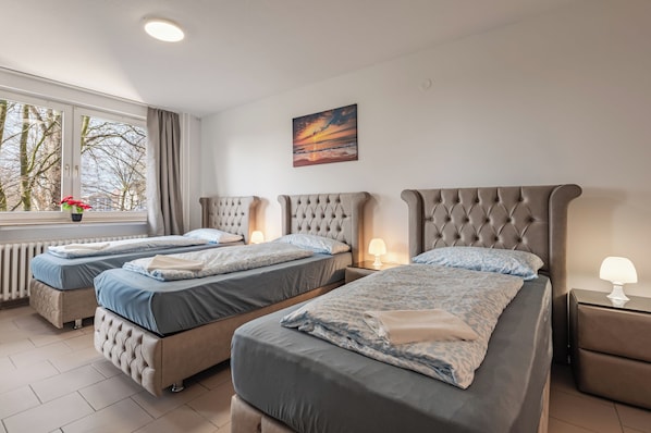 A hotel room with three beds, a window with curtains, a painting on the wall, and a radiator.