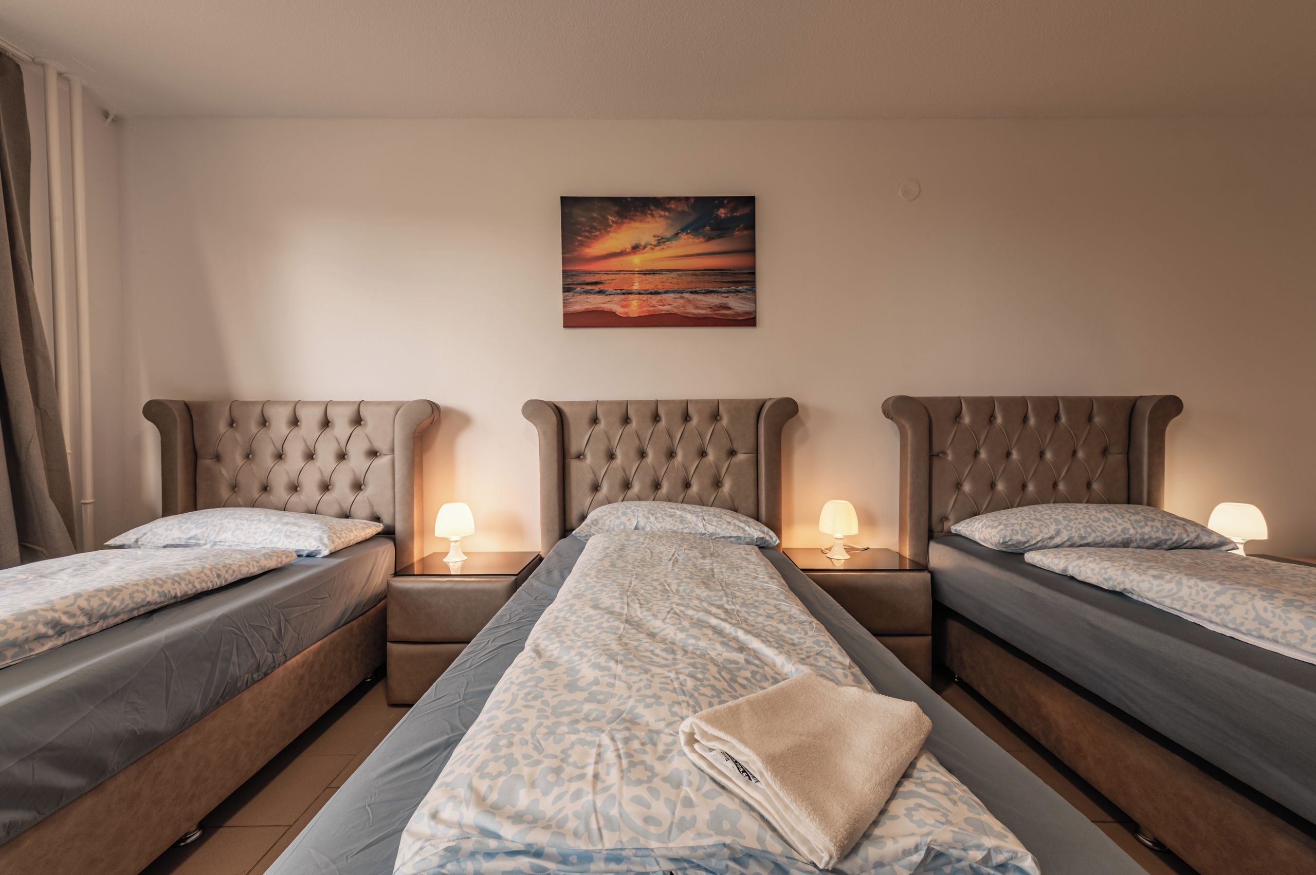 Een hotelkamer met drie eenpersoonsbedden, elk met hoofdeind, nachtkastjes en lampen. Op de muur hangt een schilderij van een zonsondergang.
