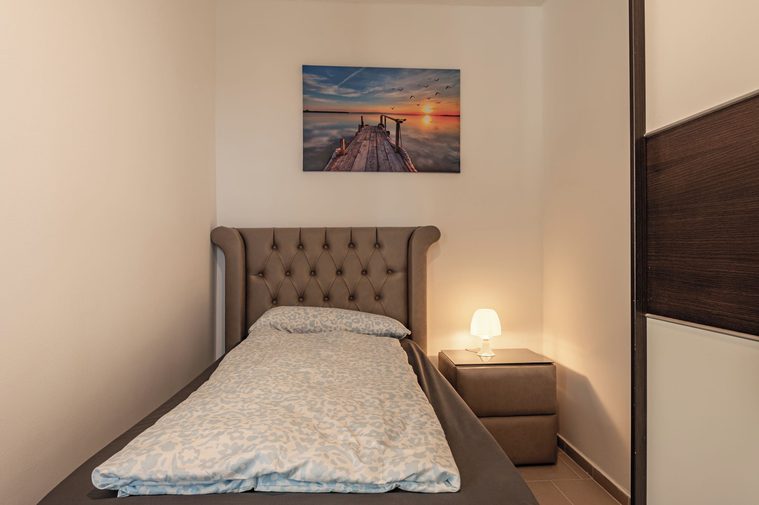 Una cama individual con cabecera, una mesita de noche con lámpara, y un cuadro colgado en la pared que muestra un atardecer sobre un muelle.