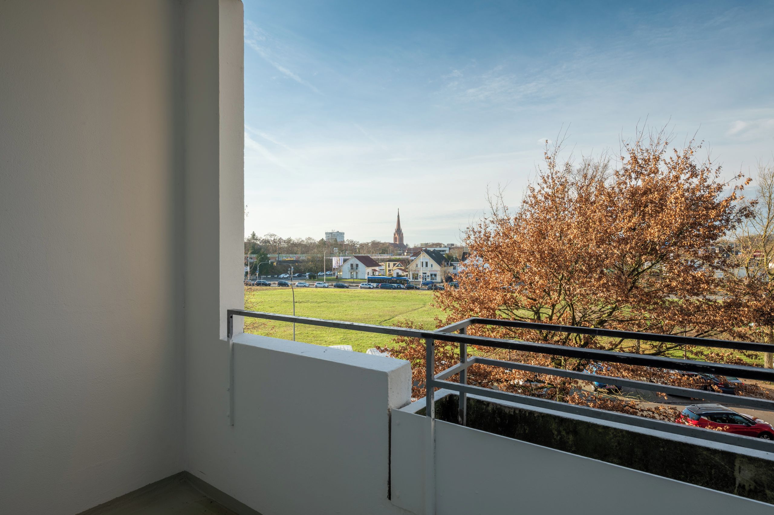 Een balkon met uitzicht op een kerktoren, een boom met herfstbladeren en een grasveld.