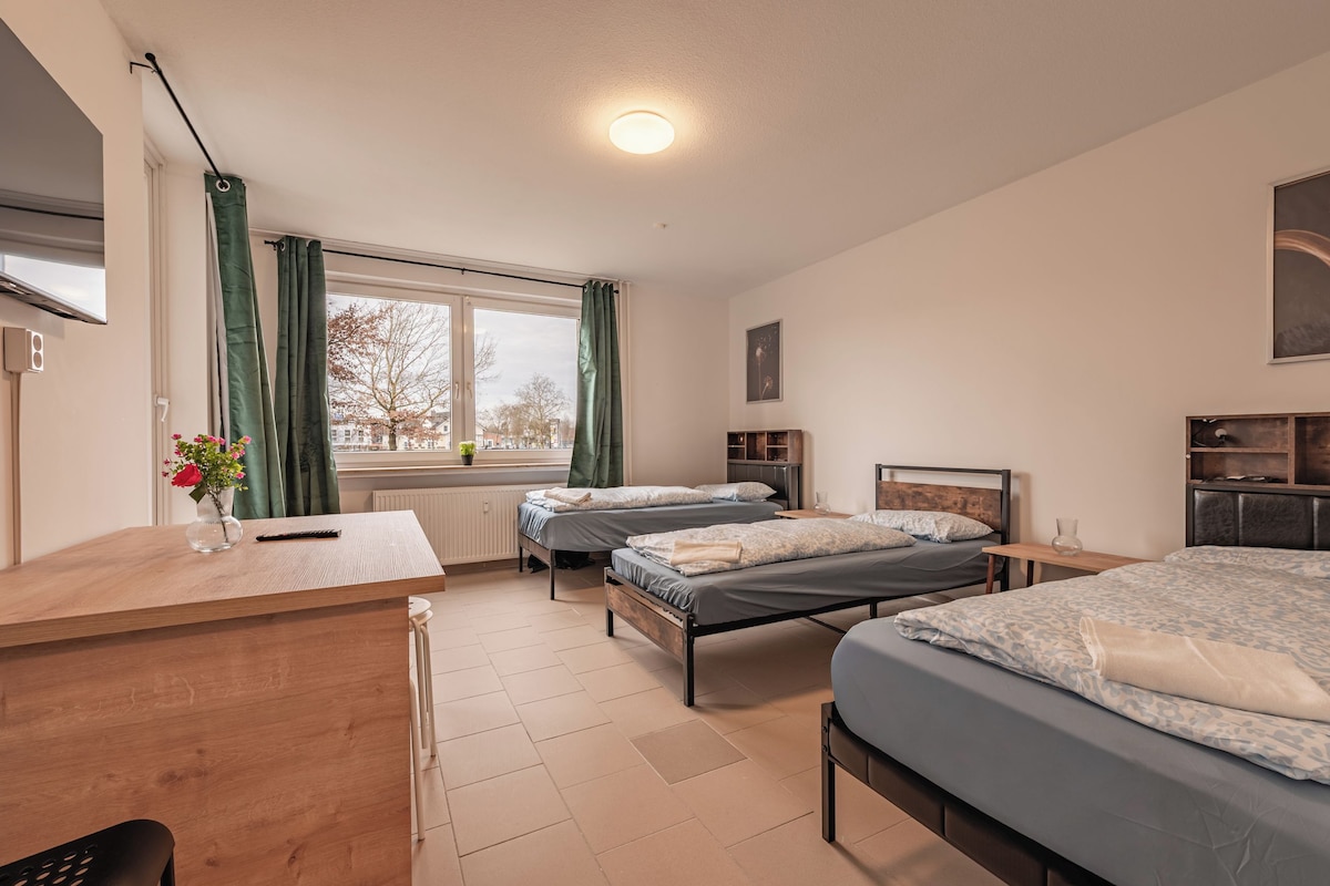 Habitació d'hotel amb dos llits, una escriptori, una finestra amb cortines, un radiador i una prestatgeria penjada a la paret.