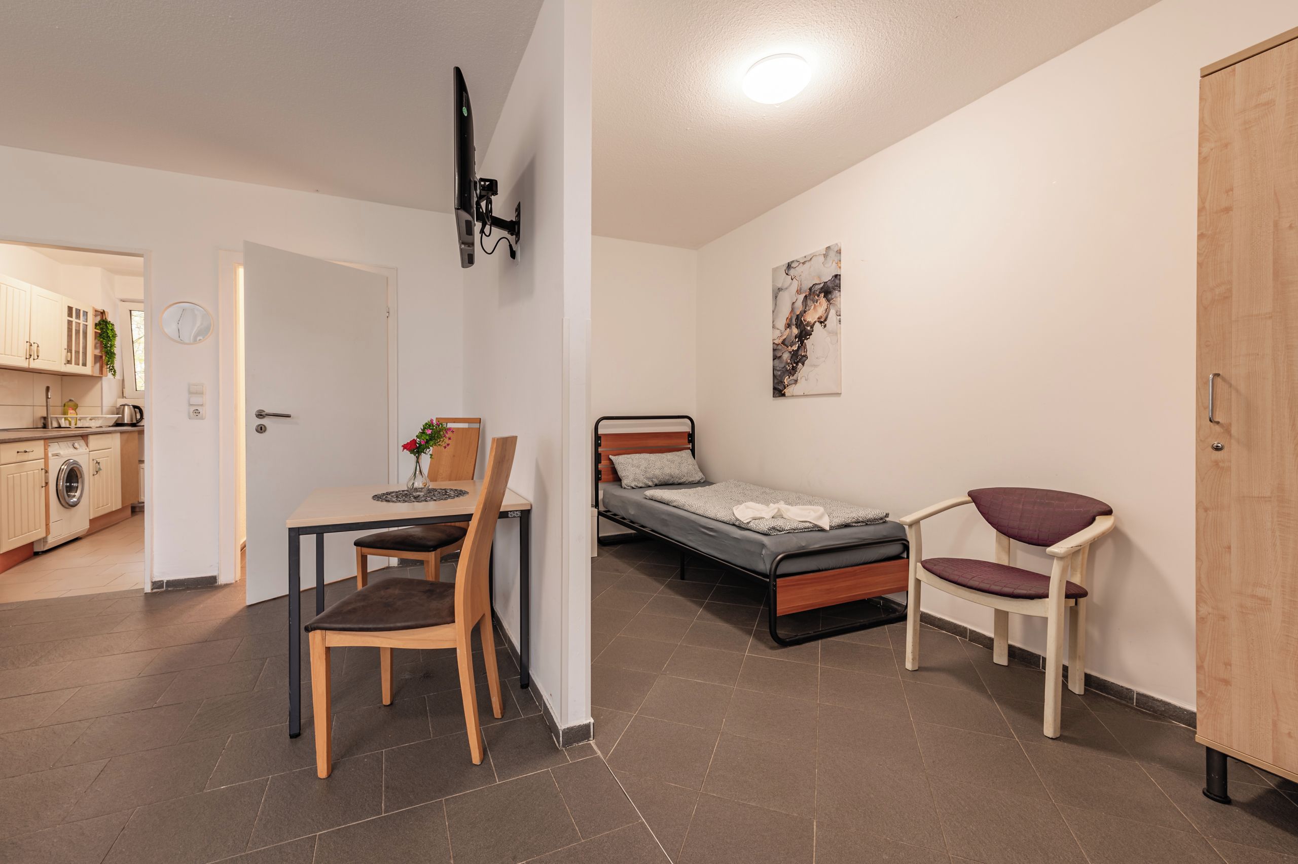 A compact room with a bed, a desk, and a chair. There is a kitchenette in the background.