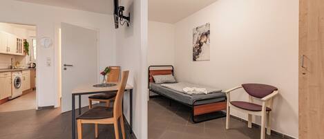 A compact room with a bed, a desk, and a chair. There is a kitchenette in the background.