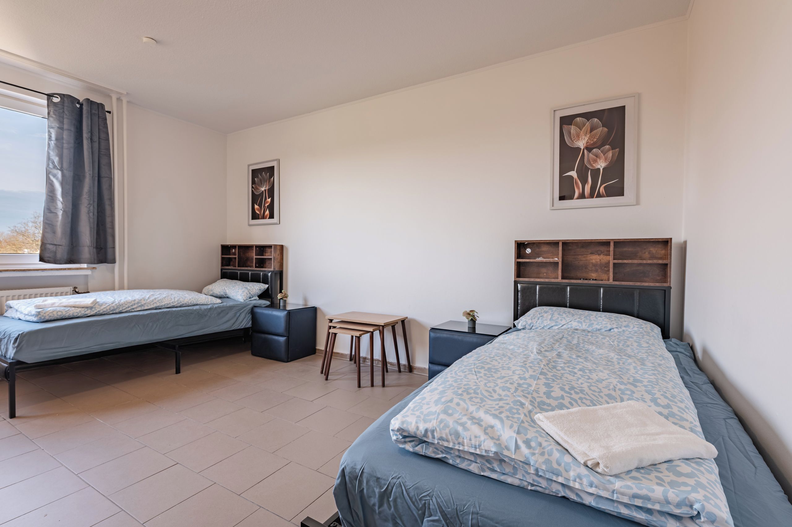 A bedroom with two beds, a small table, and a window with curtains.
