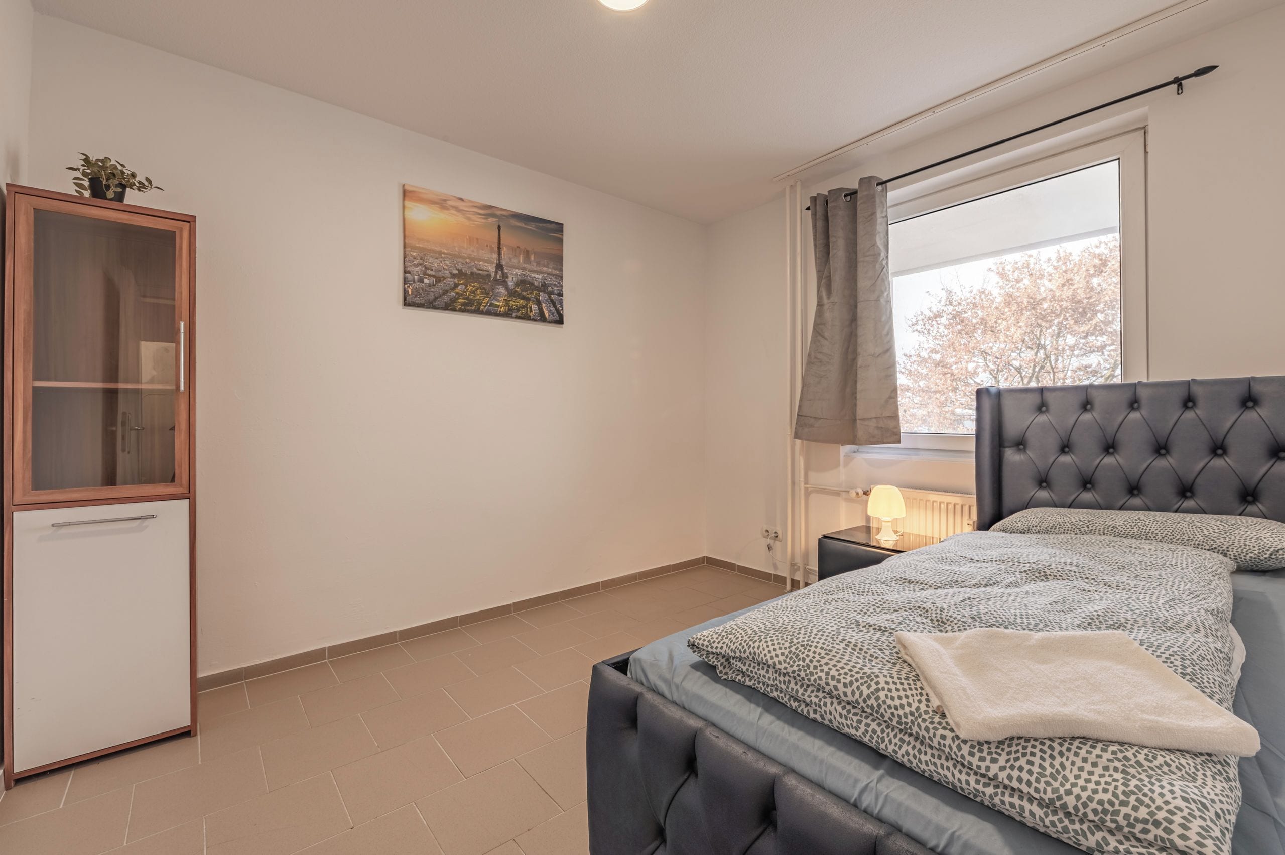 Un dormitorio con cama, mesita de noche, lámpara, ventana con cortinas, armario y un cuadro de paisaje urbano en la pared.