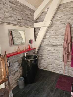 Un baño con pared de piedra, encimera roja, lavabo negro, espejo, perchero y silla de madera.