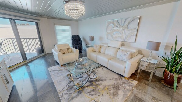 Un salon spacieux avec une table basse en verre, un canapé blanc, un lustre et un grand tableau abstrait.