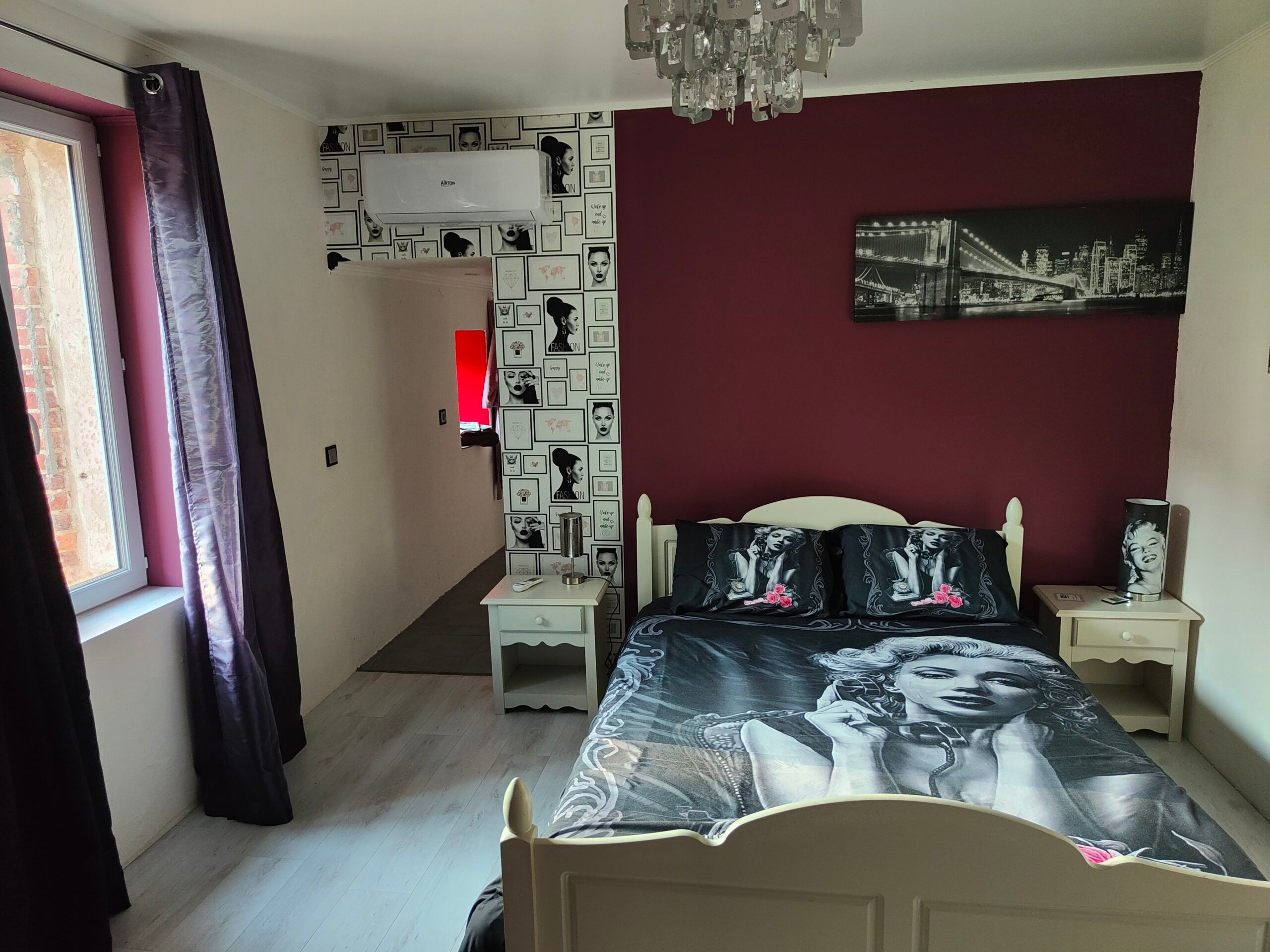 Une chambre à coucher comprenant un lit, une table de chevet, une commode, un climatiseur fixé au mur, une fenêtre avec des rideaux et un tableau représentant un paysage urbain accroché au mur.