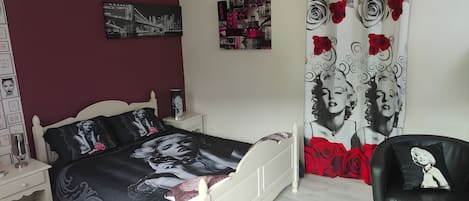 Un dormitorio con cama, mesita de noche, una silla, lámpara colgante y un cuadro de paisaje urbano en la pared.