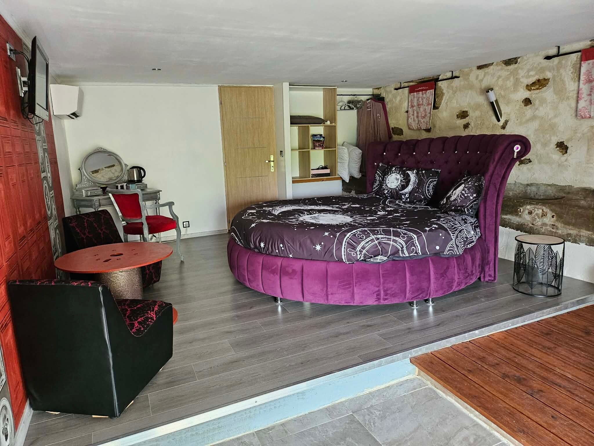 Une chambre à coucher avec un lit violet aux courbes originales, une petite table ronde, une chaise rouge et une armoire en bois.