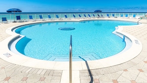 Una piscina curvada con superficie de agua azul cristalina, rodeada de sillones de descanso y sombrillas, con vista al océano.