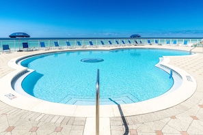 Piscina circular com dreno central, rodeada por espreguiçadeiras e guarda-sóis, e vista para o oceano.