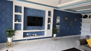 A living room with a flat-screen TV, white shelving, and potted plants. - Kijani Beach Homes Apartments Malindi (Malindi)