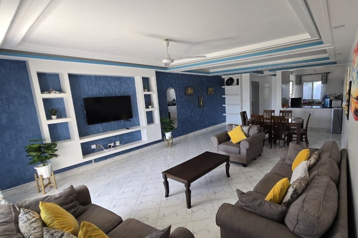 A modern living room with a flat-screen TV, sofas, a coffee table, and a dining area with chairs.