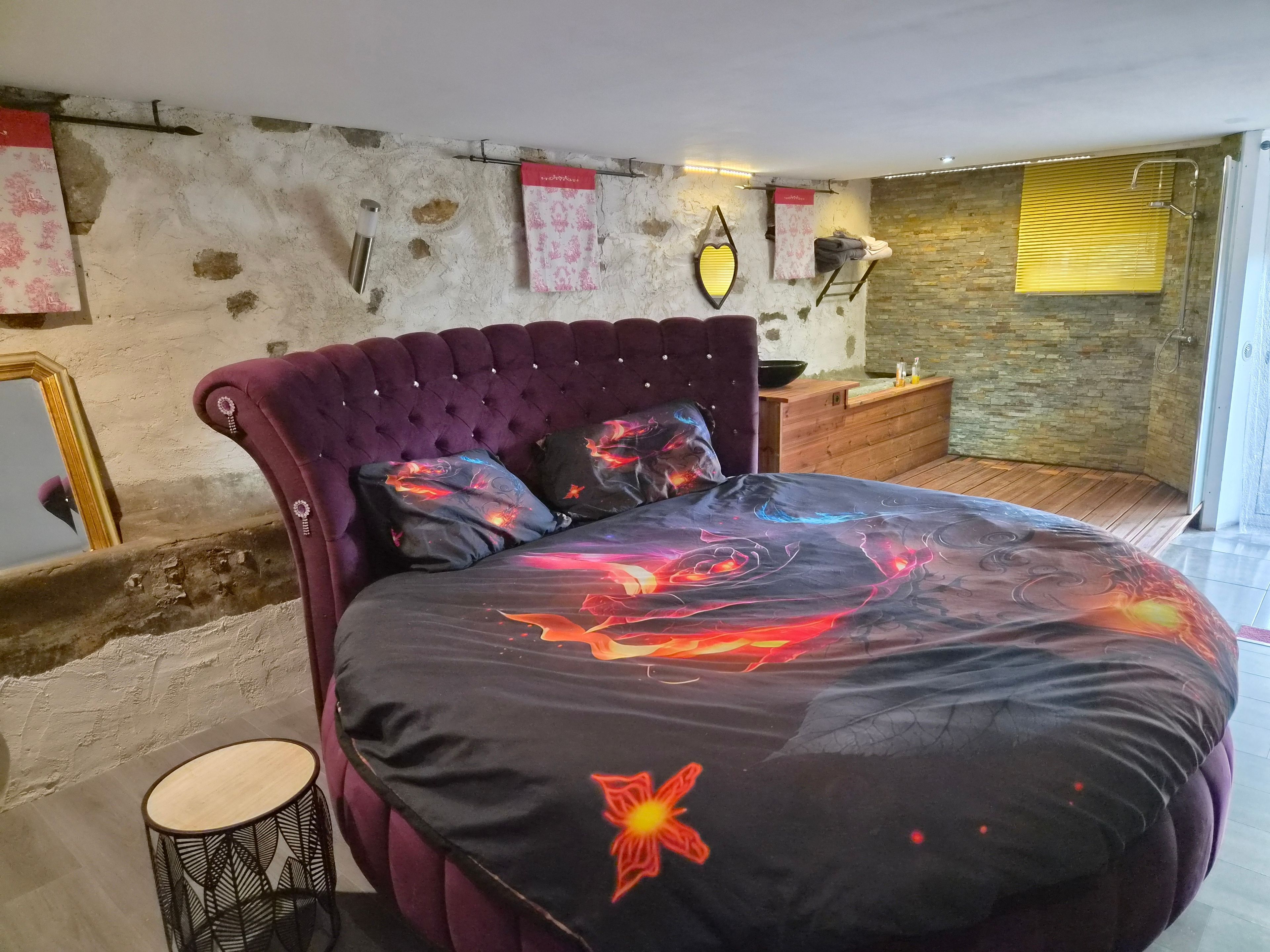 Une chambre à coucher avec une tête de lit capitonnée, un lit recouvert d’une couette sur le thème de la galaxie, une petite table ronde et une table de chevet en bois.