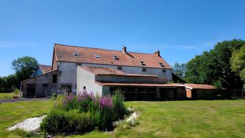 Une maison à deux étages, dotée d’un toit en pente, d’un porche à l’avant et d’un petit jardin agrémenté de fleurs violettes.