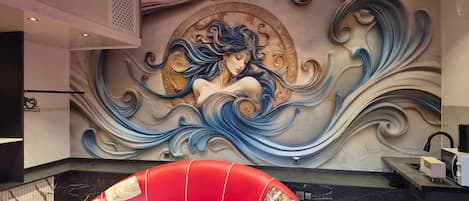 Una habitación con un mural de una mujer en la pared, un sillón tapizado rojo y una encimera de mármol negro.