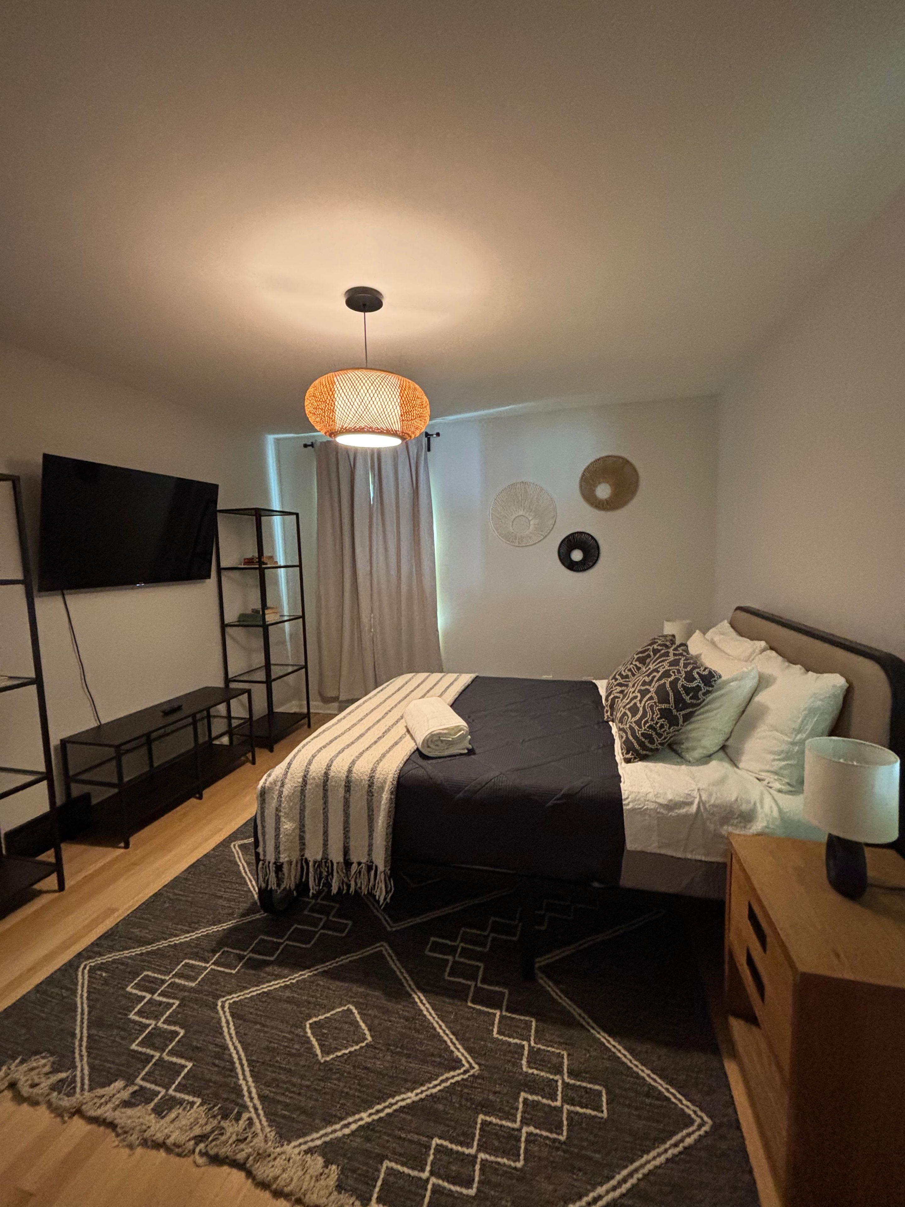 A bedroom with a bed, a TV, a shelf, a lamp, and a patterned rug.