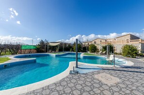 Une piscine au design incurvé, entourée d’une terrasse en pierre et bordée d’un bâtiment à la façade en pierre.