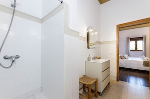 Een badkamer met witte tegels, een douche, een wastafel met spiegel en een houten kruk.