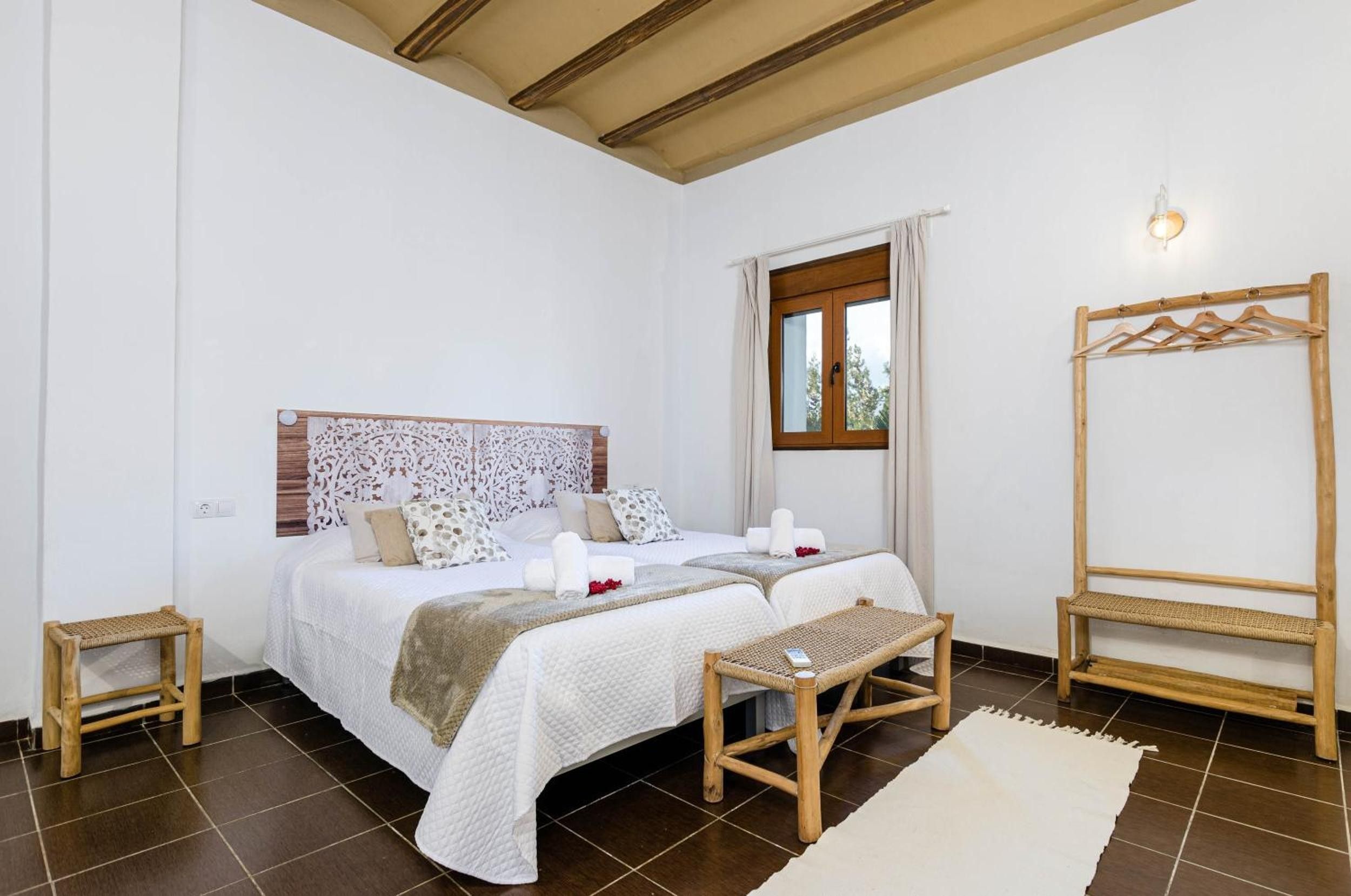 A bedroom with a large bed, two bedside tables, a wooden bench, a hanging clothes rack, and a window with curtains.
