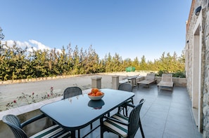 Um alpendre ao ar livre com mesa e cadeiras, uma cesta de frutas e vista para árvores e um edifício.