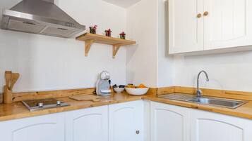 Una cocina con armarios blancos, extractor de acero inoxidable, encimera de madera, cafetera, un tazón de fruta y un lavamanos.