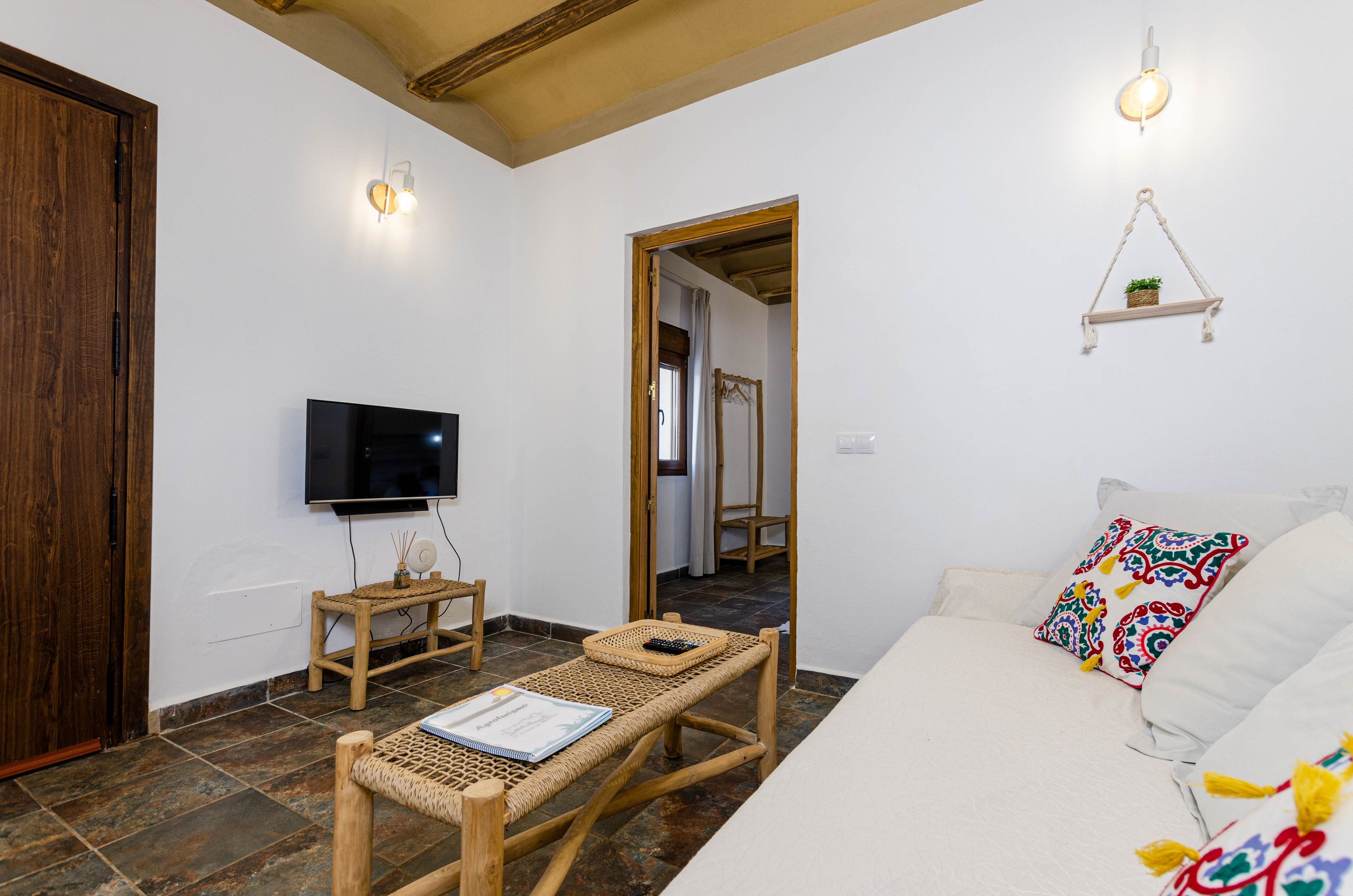 Una habitación con piso de madera, un sofá blanco con almohadones de colores, un televisor en la pared, una mesita con un libro y una planta colgante.