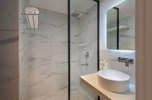 A modern bathroom with a marble wall, a glass-enclosed shower, a white sink, and a mirror. - ONE Atmosphere Suites - Corner Walk (Gurugram)