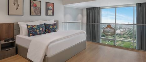 A modern hotel room with a large bed, a nightstand, a window with a view, and artwork on the wall.