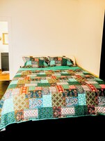 Una cama con colcha y almohadas de colores, una pared blanca y una puerta.